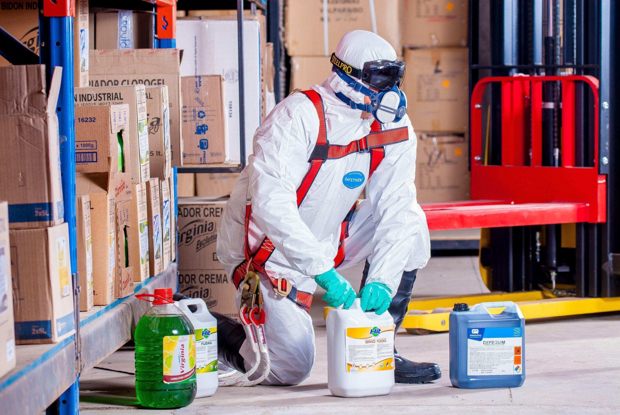 Person wearing a white hazmat suit carrying toxic chemicals in an industrial setting.